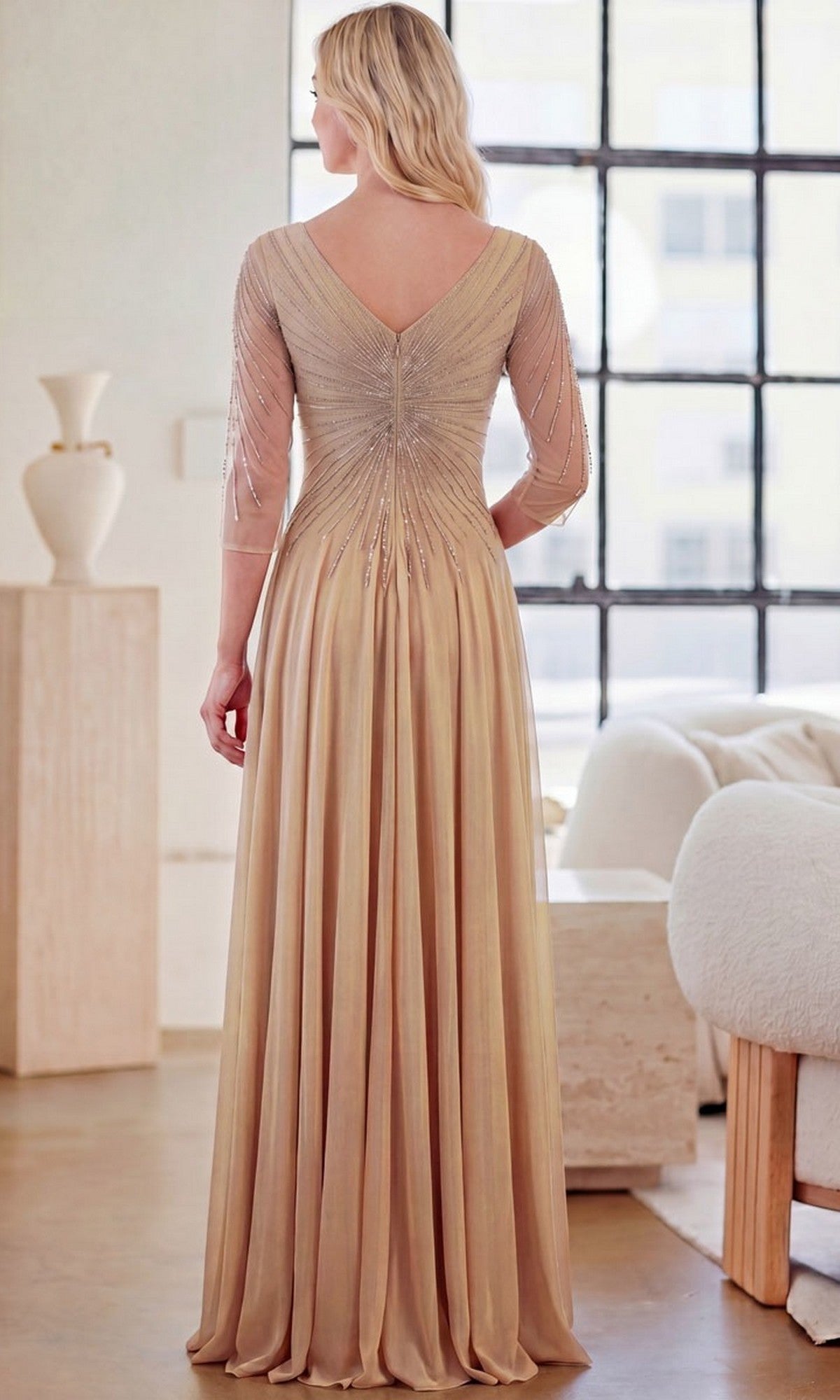 A woman stands indoors wearing the Long Formal Dress: Amelia Couture KF1501, featuring a beaded bodice, V-back, and an elegant A-line skirt.